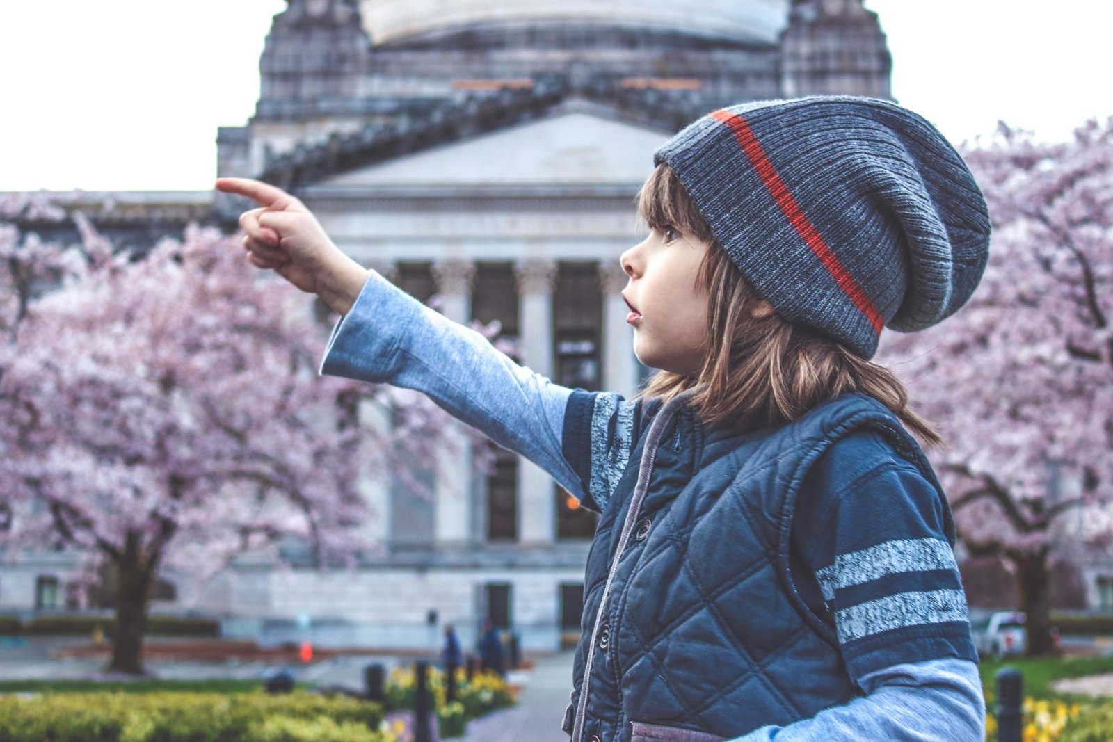 A young girl in a beanie points towards cherry blossoms in front of a historic building.