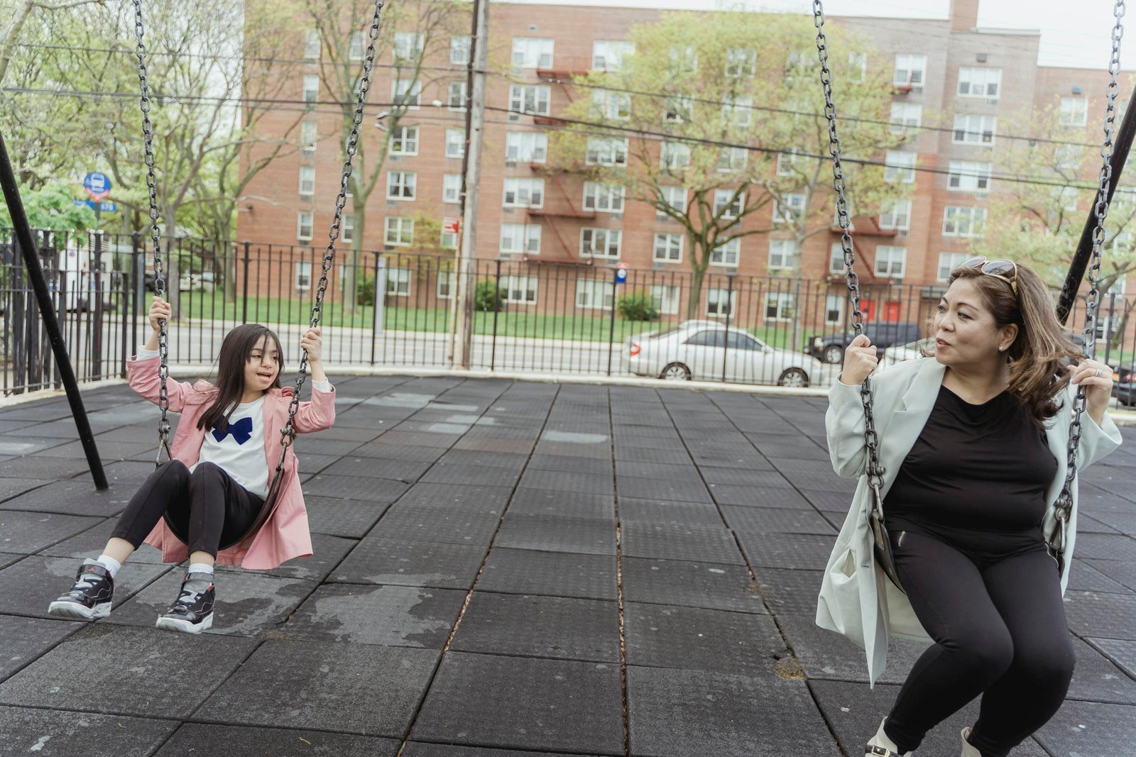 Mother and daughter enjoy quality time swinging together at the playground. Urban setting with spring vibes.