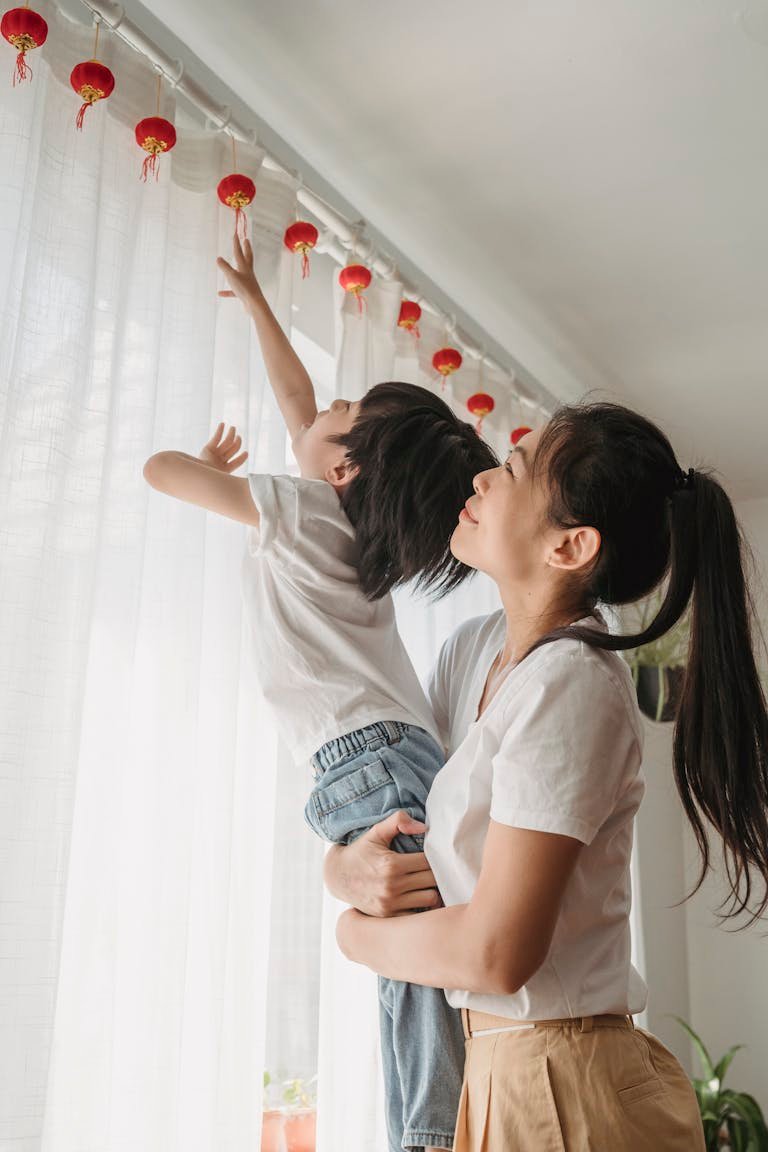 Asian mother holding son as he reaches for red lanterns in a warm indoor setting.