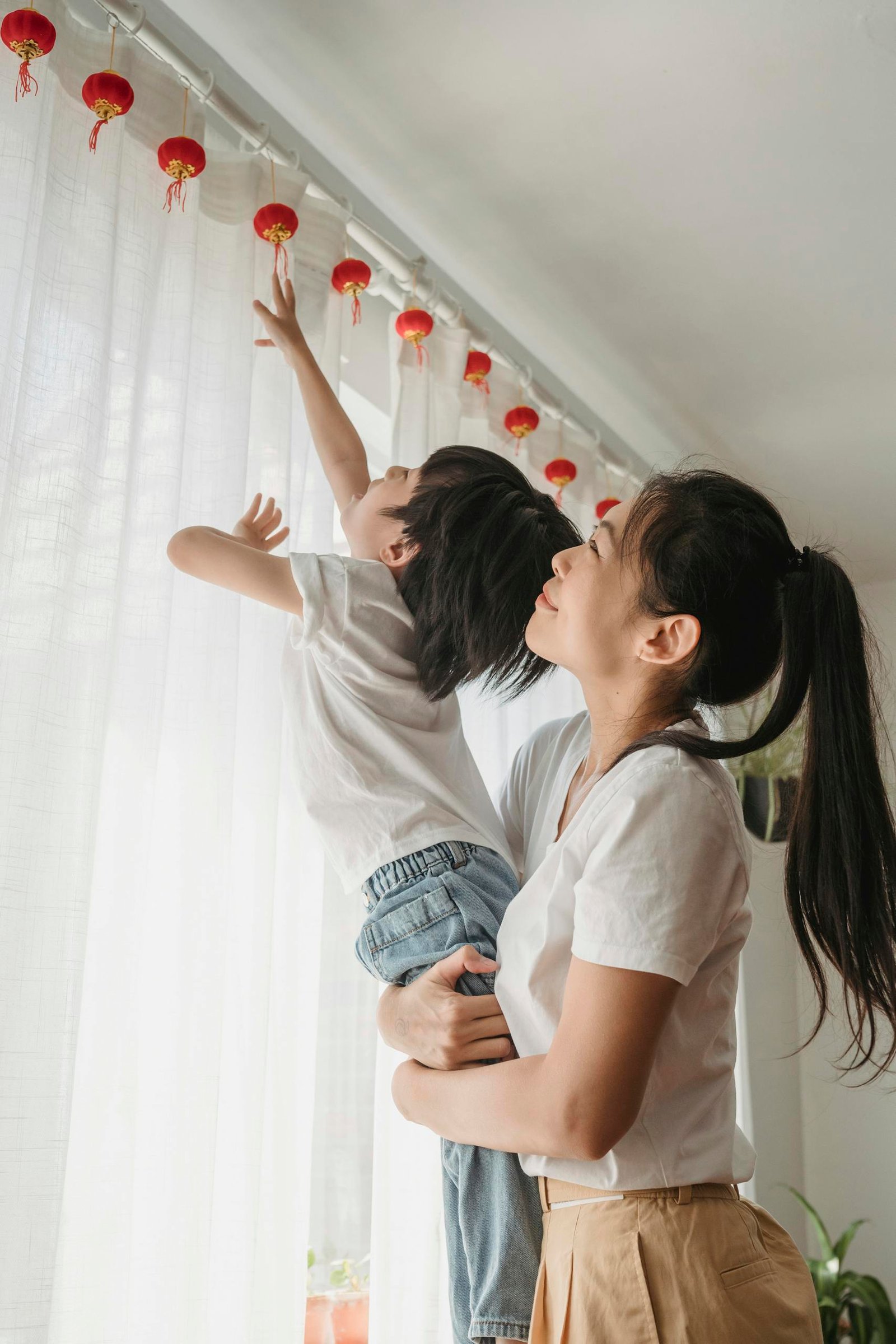 Asian mother holding son as he reaches for red lanterns in a warm indoor setting.